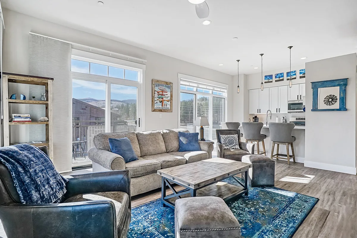 A spacious, airy living area with a comfortable sofa, coffee table, and a rug facing large windows that offer sweeping mountain views. The adjoining kitchen area is visible, highlighting the seamless open-plan design. Soft, natural light streams in, and the ambiance is serene and inviting.