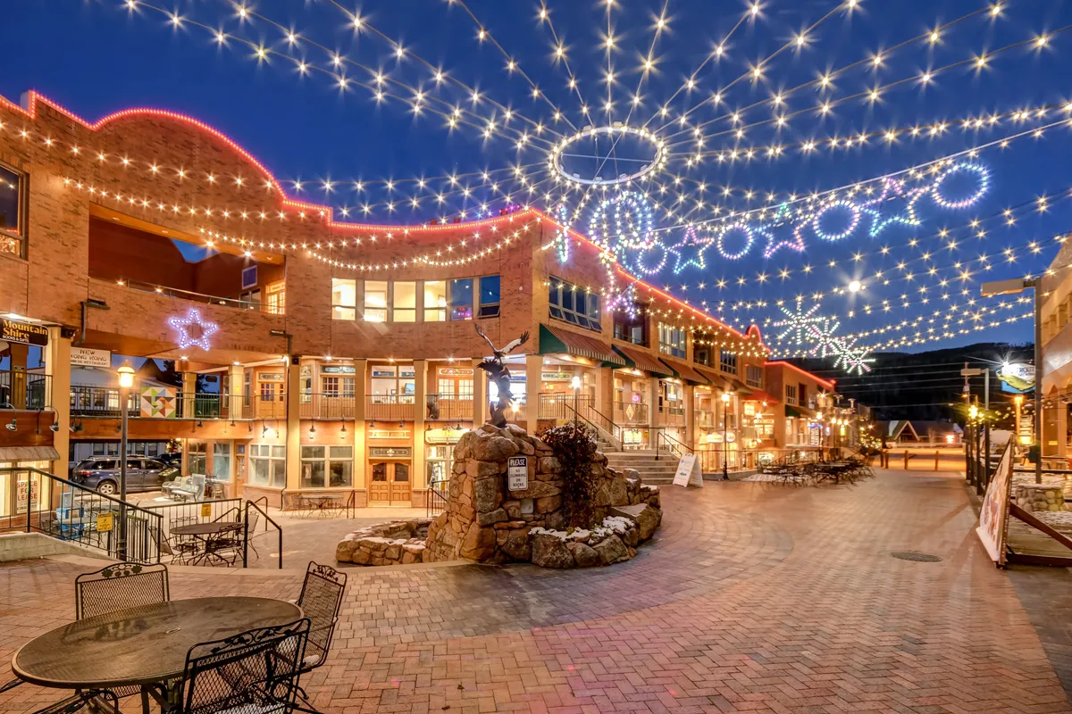 An inviting exterior scene illuminated by string lights and decorated with hanging stars. The view includes a stone fountain and a charming brick pathway surrounded by people engaging in social gatherings and enjoying the lively ambiance. The scene suggests close proximity to shops and restaurants, capturing a festive downtown atmosphere.