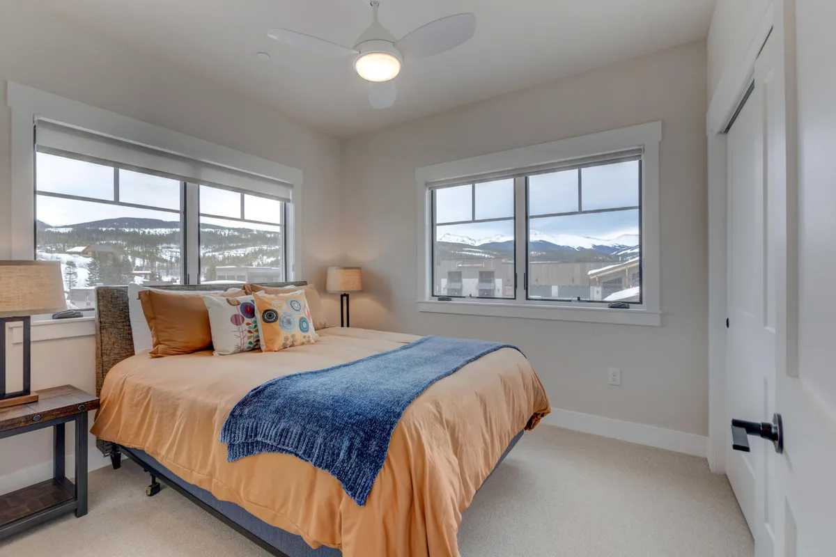 A serene bedroom with a plush bed adorned with decorative pillows, large windows offering mountain views, and a nightstand with a lamp. The natural light and inviting bedding create an atmosphere of comfort and relaxation—perfect for a restful retreat.