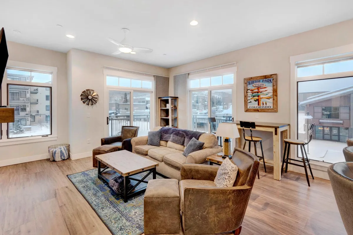 A cozy living room featuring a comfortable sofa and a wooden coffee table, framed by large windows that let in natural light and provide views of the outdoors. Decorative wall art adds a touch of warmth, and the space feels inviting for relaxation or group gatherings.