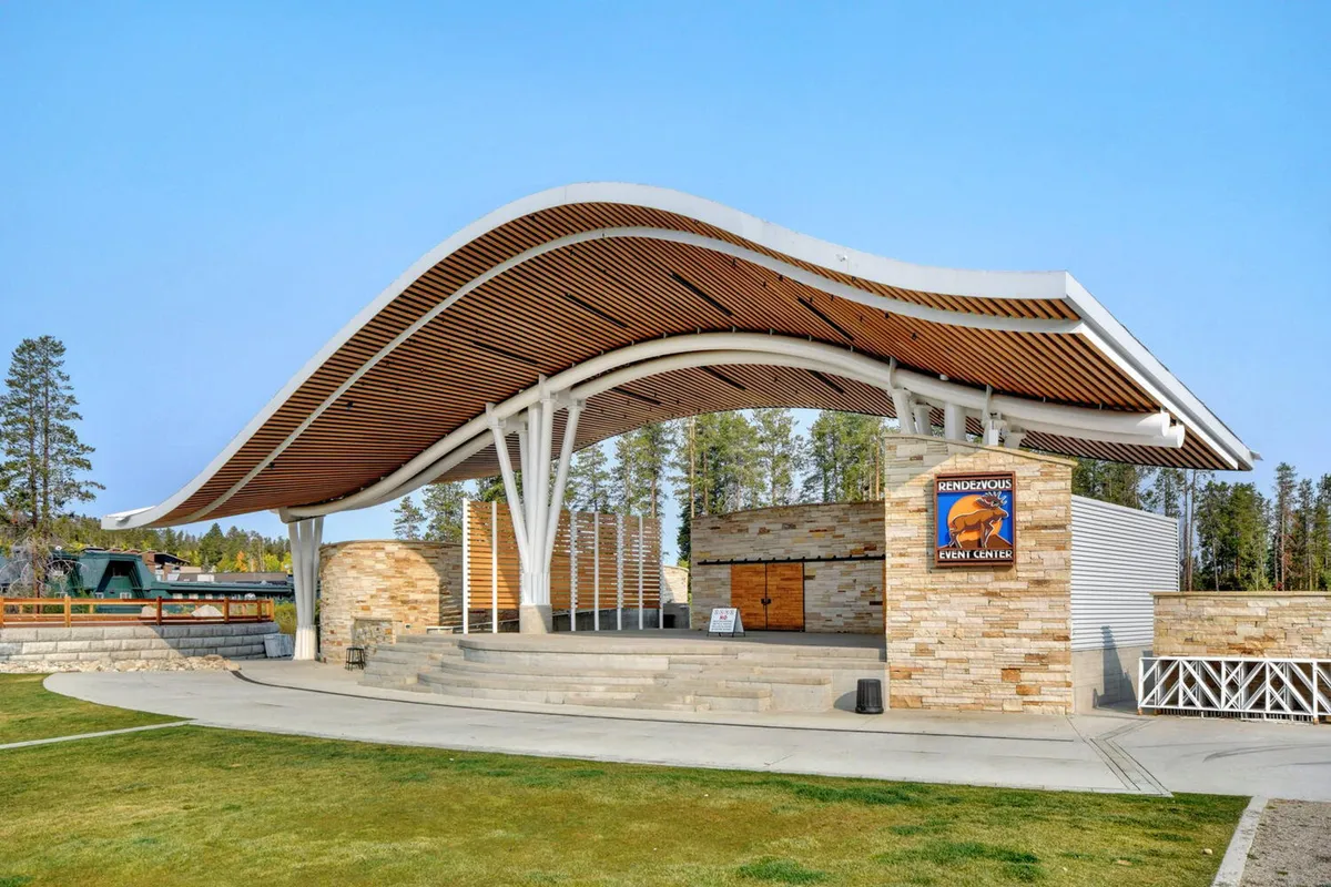 An exterior shot of the Hideaway Park Amphitheater, featuring a modern curved roof structure, stone walls, and an open stage area, set up for outdoor concerts and community events. The scene suggests the excitement and community spirit of open-air gatherings.