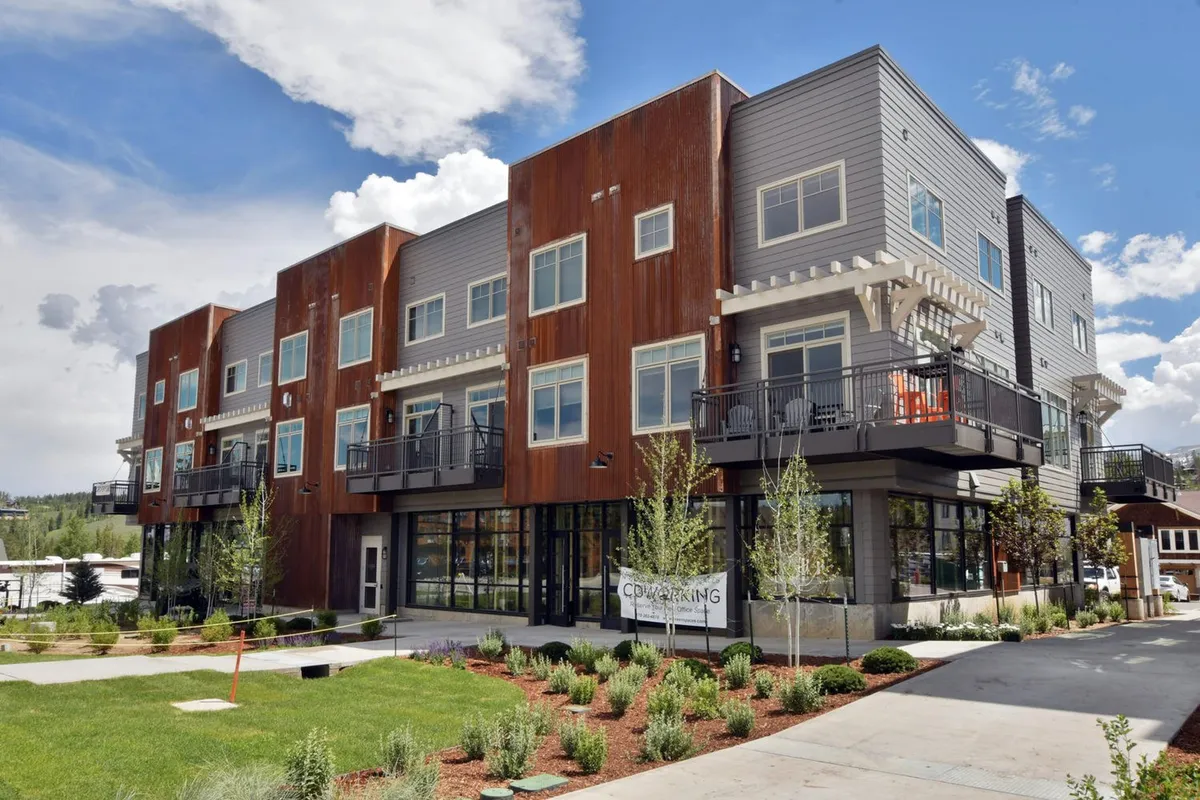 A contemporary exterior view of the Luxury Loft building, featuring modern architecture with large balconies and expansive windows. Lush green landscaping surrounds the property, while people relax on balconies and mingle in the courtyard, suggesting a vibrant, social atmosphere just steps from the excitement of downtown and resort activities.