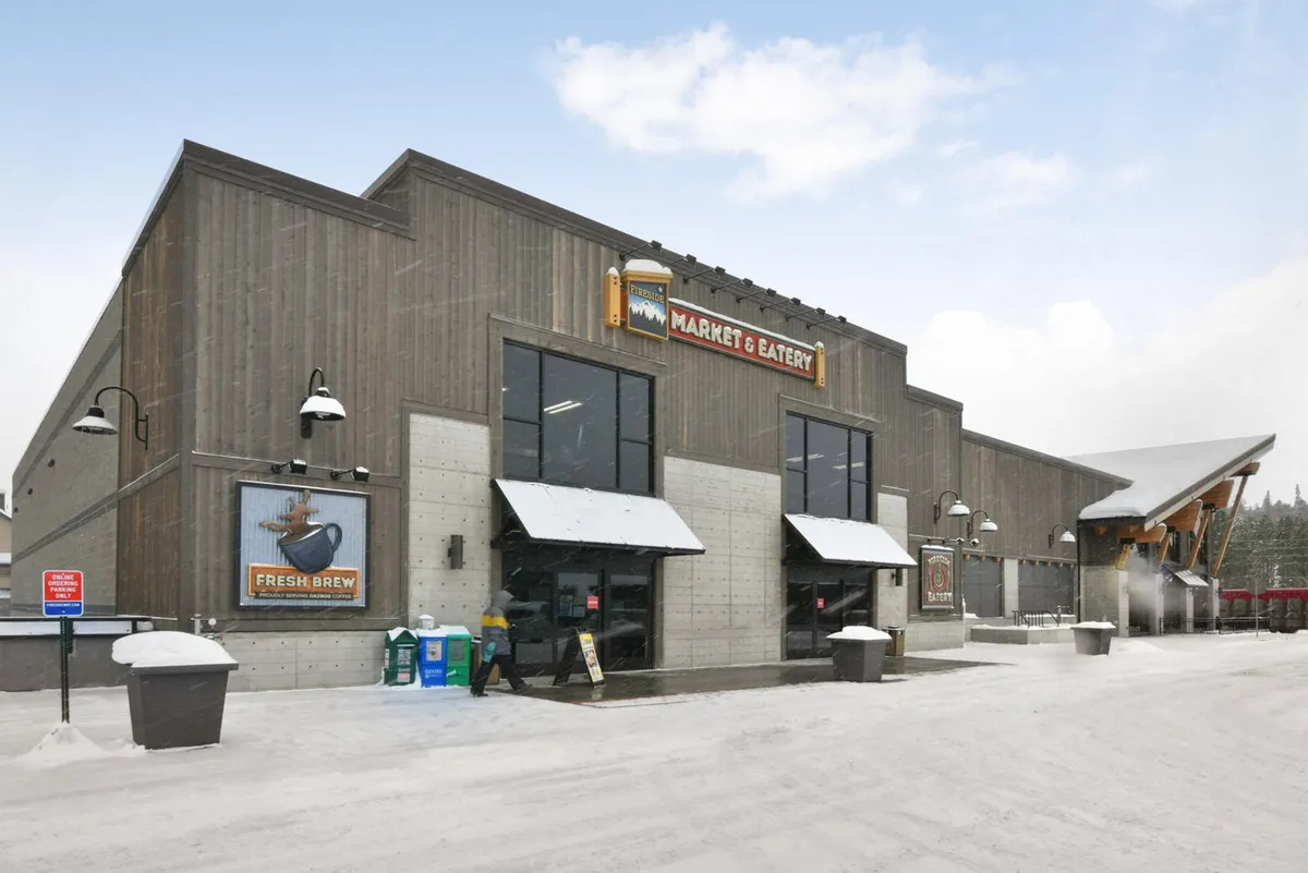 A cozy, snow-dusted exterior view of the Market & Eatery, featuring a modern wooden structure, inviting signage, outdoor seating, and a welcoming atmosphere. The scene suggests warmth and togetherness after a day of exploration.