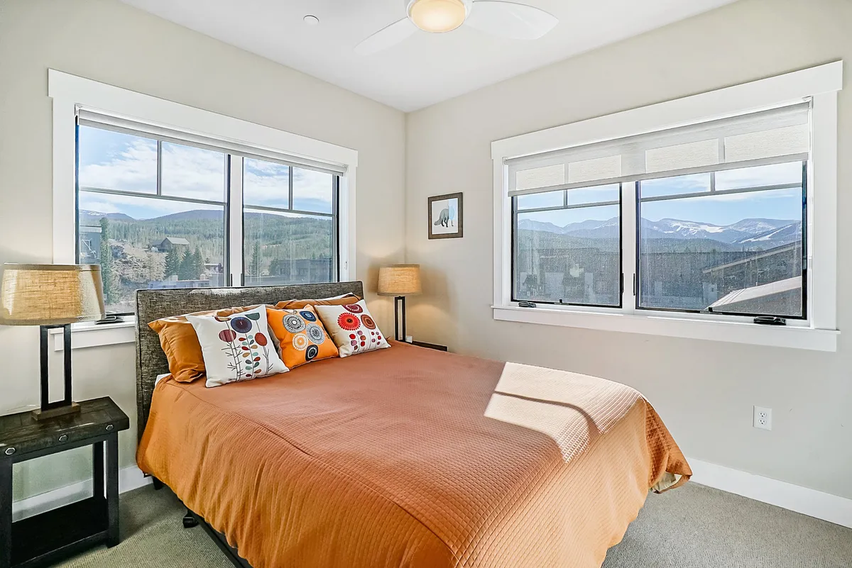 A cozy bedroom scene featuring a bed with an orange coverlet and decorative pillows. Large windows frame stunning mountain vistas, suggesting a tranquil retreat. The room exudes comfort and relaxation, hinting at premium bedding and thoughtful touches.